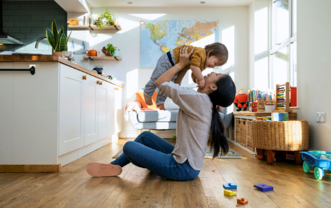 Erwachsene Person spielt mit Kleinkind im sonnigen Wohnzimmer mit Holzboden und Weltkarte an der Wand. | Quelle: iStock
